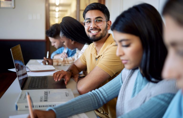A man touching his laptop smiling while looking at the camera and others in the background writing on a book